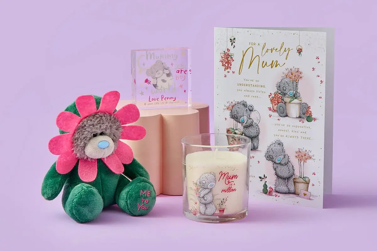 A cute Mother’s Day gift set featuring a flower‑costumed teddy bear, a scented candle with a bear illustration, a small personalised acrylic block, and a floral card, all arranged on a pastel purple background.